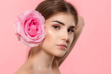 Pembe arka plan üzerinde izole yüz yakın pembe gül çiçeği tutan kameraya bakan güzel bir genç kızın yakın çekim portre.