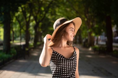 Güzel genç esmer kadın siyah bir elbise ve geniş kapaklı bir şapka giymiş sıcak yaz günü güneş ışığı zevk sırasında bir parkta yürüyüşe çıkar.