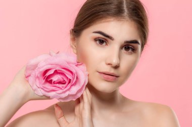 Pembe arka plan üzerinde izole yüz yakın pembe gül çiçeği tutan kameraya bakan güzel bir genç kızın yakın çekim portre.
