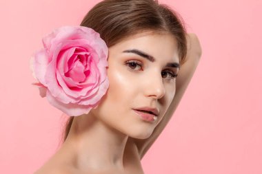 Pembe arka plan üzerinde izole yüz yakın pembe gül çiçeği tutan kameraya bakan güzel bir genç kızın yakın çekim portre.
