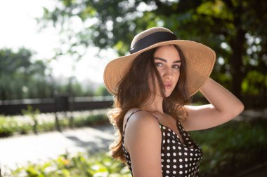 Siyah bir elbise ve geniş kapaklı bir şapka giymiş güzel bir genç esmer kadının yakın çekim porttrait sıcak yaz günü güneş ışığı zevk sırasında bir parkta bir yürüyüş alır.