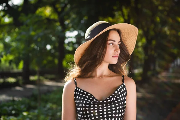 Retrato de cerca de una hermosa joven morena vestida con un vestido negro y un sombrero con solapas anchas da un paseo en un parque durante el cálido día de verano disfrutando de la luz del sol . — Foto de Stock