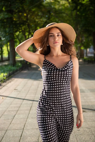 Beautiful young brunette woman dressed in a black dress and a hat with wide flaps takes a walk in a park during warm summer day enjoying sunlight. — Stock Photo, Image