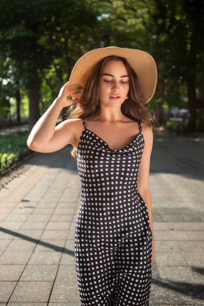 Vacker ung brunett kvinna klädd i en svart klänning och en hatt med breda flikar tar en promenad i en Park under varm sommardag njuter av solljus. — Stockfoto