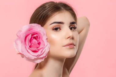 Pembe arka plan üzerinde izole yüz yakın pembe gül çiçeği tutan kameraya bakan güzel bir genç kızın yakın çekim portre.