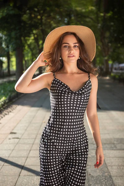 Beautiful young brunette woman dressed in a black dress and a hat with wide flaps takes a walk in a park during warm summer day enjoying sunlight. — Stock Photo, Image