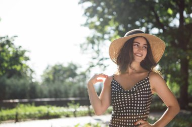 Güzel genç esmer kadın siyah bir elbise ve geniş kapaklı bir şapka giymiş sıcak yaz günü güneş ışığı zevk sırasında bir parkta yürüyüşe çıkar.