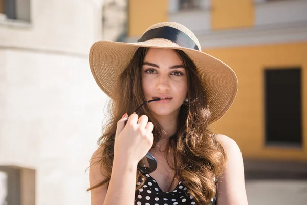 Chica bonita en un vestido negro y un sombrero con amplias aletas sonrisas sosteniendo gafas de sol en los dientes disfrutando de caminar por la ciudad europea durante el soleado día de verano . — Foto de Stock