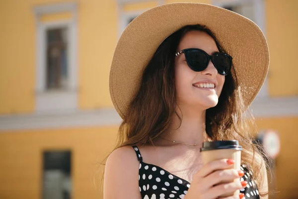 Chica bonita en un vestido negro, sombrero con solapas anchas y gafas de sol sostiene la taza de papel desechable de café caliente disfrutando de caminar por la ciudad europea durante el soleado día de verano . — Foto de Stock