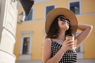 Siyah elbiseli güzel kız, geniş kapaklı şapka ve güneş gözlüğü ile güneşli yaz gününde Avrupa kasabasında yürümenin keyfini çıkarın sıcak kahve tek kullanımlık kağıt fincan tutar.
