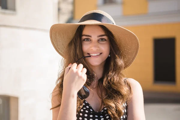 Chica bonita en un vestido negro y un sombrero con amplias aletas sonrisas sosteniendo gafas de sol en los dientes disfrutando de caminar por la ciudad europea durante el soleado día de verano . — Foto de Stock