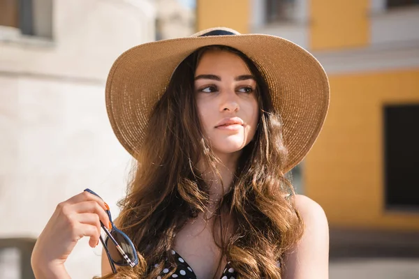 Chica bonita en un vestido negro y un sombrero con amplias aletas sonrisas sosteniendo gafas de sol disfrutando de caminar por la ciudad europea durante el soleado día de verano . — Foto de Stock