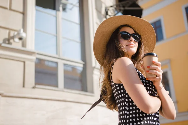 Chica bonita en un vestido negro, sombrero con solapas anchas y gafas de sol sostiene la taza de papel desechable de café caliente disfrutando de caminar por la ciudad europea durante el soleado día de verano . — Foto de Stock