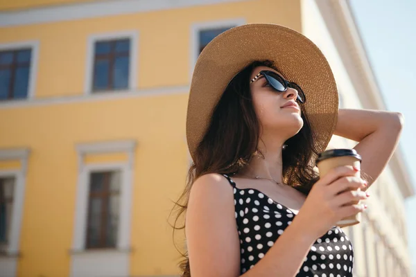 Chica bonita en un vestido negro, sombrero con solapas anchas y gafas de sol sostiene la taza de papel desechable de café caliente disfrutando de caminar por la ciudad europea durante el soleado día de verano . — Foto de Stock
