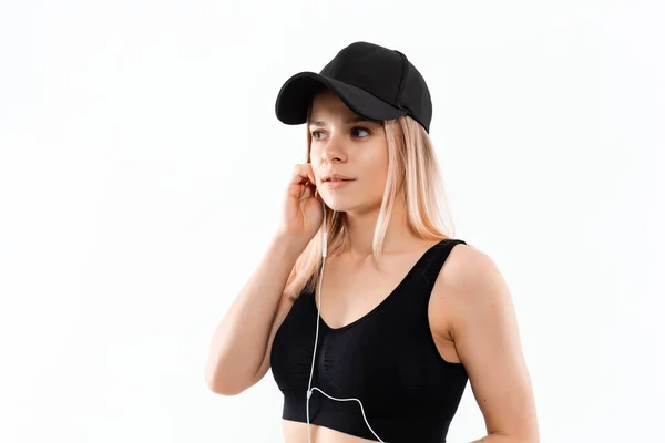 Joven mujer rubia deportiva en una ropa deportiva negra con relojes inteligentes escucha música durante el entrenamiento de pie sobre fondo blanco . — Foto de Stock