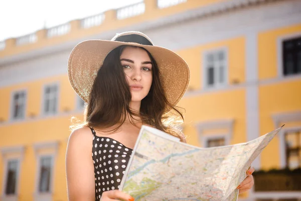 Chica bonita en un vestido negro y sombrero con solapas anchas sostiene mapa averiguar la ruta durante el recorrido turístico mientras viaja por el casco antiguo europeo . — Foto de Stock