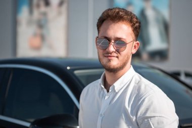 Park halindeki arabanın yanında duran beyaz gömlek li ve güneş gözlüklü genç başarılı işadamı.