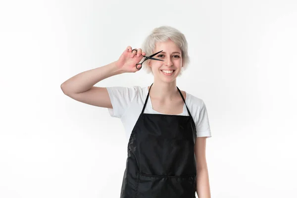 Hübsche junge Friseurin in weißem T-Shirt und schwarzer Schürze mit schwarzer Schere vor dem Gesicht, bereit für die Arbeit, isoliert vor weißem Hintergrund stehend. — Stockfoto