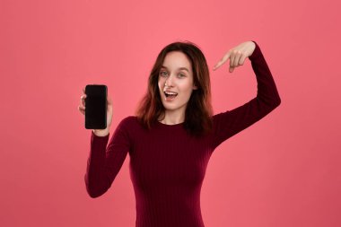 Heyecanlı güzel esmer kız kameraya bakarak koyu pembe arka plan üzerinde dururken bir parmak ile bir cep telefonu ekranına işaret.