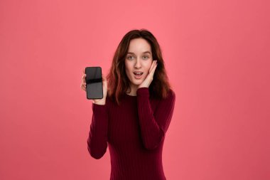 Heyecanlı güzel esmer kız kameraya bakarak koyu pembe bir arka plan üzerinde dururken bir cep telefonu ekran gösteren.