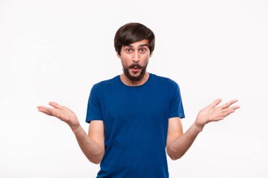Sakallı mavi gömlekli ve çoğu ağrılı, beyaz arka plan üzerinde izole ayakta duran şaşıran omuzlarını silkme iyi görünümlü brunet adam. Duygu ve şok jest.