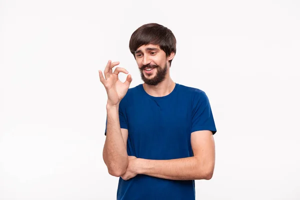 Knappe Brunet bebaarde man met snorren in een blauw overhemd aarzelend tonen gebaar van OK teken staande geïsoleerd over witte achtergrond. OK gebaar. — Stockfoto