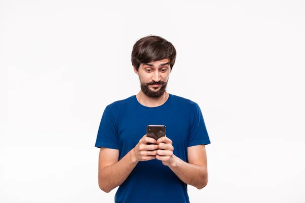 Good looking brunet man in a blue shirt with beard and mostaches shocked looking at the screen of a smartphone standing isolated over white background. Emotion and gesture of surprise. — Stock Photo, Image