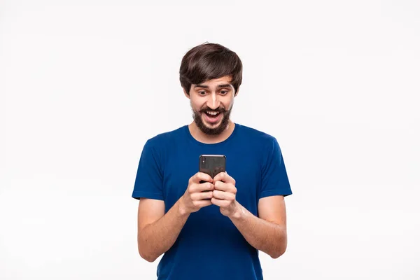 Good looking brunet man in a blue shirt with beard and mostaches surprised looking at the screen of a smartphone standing isolated over white background. Emotion and gesture of shock. — Stock Photo, Image