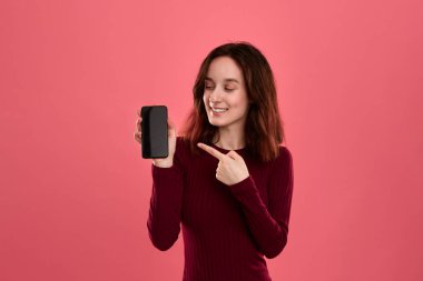 Heyecanlı güzel esmer kız koyu pembe arka plan üzerinde dururken bir parmak ile bir cep telefonu ekranında işaret .
