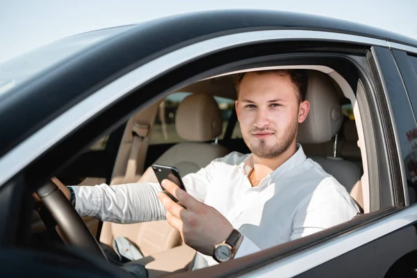 Joven hombre de negocios sonriendo mirando el teléfono móvil mientras conduce un coche . — Foto de Stock