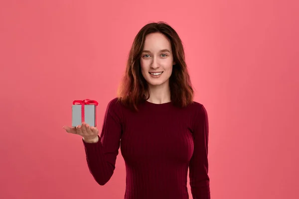 Excited pretty brunette girl holding present box with a ribbon standing isolated on a dark pink background and smiling at the camera. Celebrating special event. — Stock Photo, Image