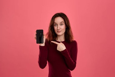 Heyecanlı güzel esmer kız kameraya bakarak koyu pembe arka plan üzerinde dururken bir parmak ile bir cep telefonu ekranına işaret.