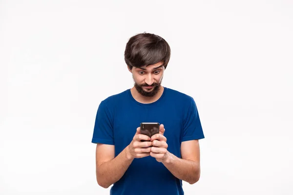 Good looking brunet man in a blue shirt with beard and mostaches shocked holding smartphone looking at the camera standing isolated over white background. — Stock Photo, Image