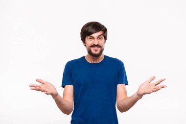 Sakallı mavi gömlekli iyi görünümlü brunet adam ve çoğu beyaz arka plan üzerinde izole ayakta şaşırarak omuzlarını silkme bağırıyor.