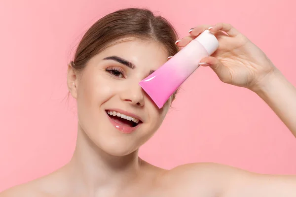 Güzel kız cildi nemlendirmek için krem tüp tutan. Model pembe arka plan üzerinde izole kamera bakar. Güzellik ve sağlık tedavisi kavramı.