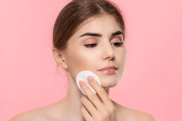 Beautiful model applyes cosmetic product to skin with white cotton sponge. Model looks at the camera isolated over pink background.