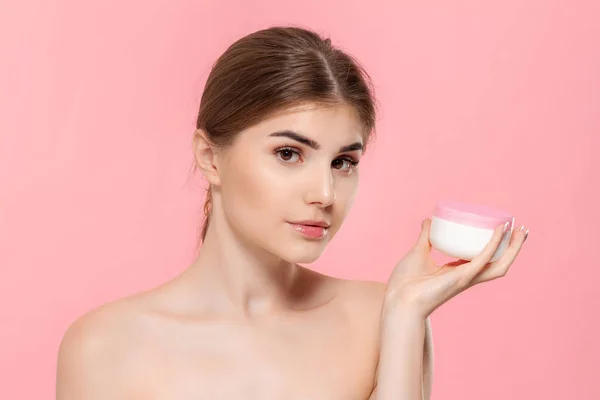 美丽的女孩拿着奶油管来滋润皮肤。模型查看在粉红色背景上隔离的摄像机。美容与健康治疗的概念. — 图库照片