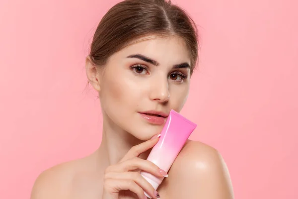 Güzel kız cildi nemlendirmek için krem tüp tutan. Model pembe arka plan üzerinde izole kamera bakar. Güzellik ve sağlık tedavisi kavramı.