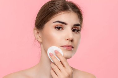 Güzel model beyaz pamuk sünger ile cilde kozmetik ürün uygular. Model pembe arka plan üzerinde izole kamera bakar.