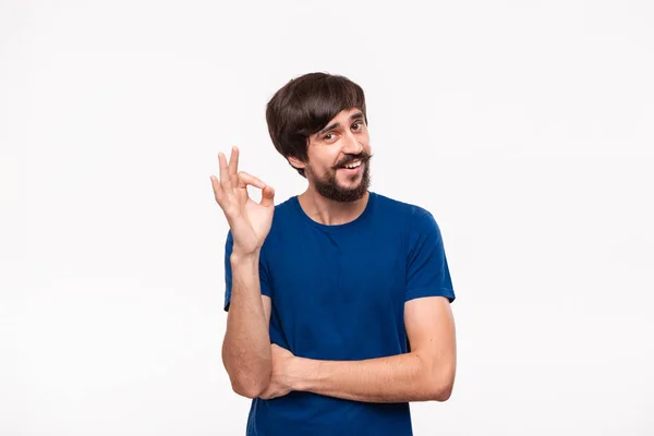 Bello uomo bruno barbuto con i baffi in una camicia blu che mostra gesto di OK segno in piedi isolato su sfondo bianco . — Foto Stock