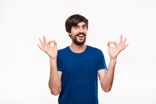 Bello uomo bruno barbuto con i baffi in una camicia blu che mostra gesto di OK segno con due mani in piedi isolato su sfondo bianco . — Foto Stock