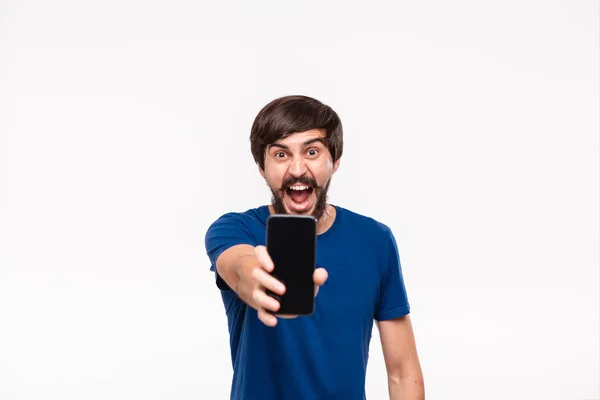 Hombre morena bien parecido en una camisa azul con barba y bigotes mostrando la pantalla del teléfono inteligente en la cámara de pie aislado sobre fondo blanco . — Foto de Stock