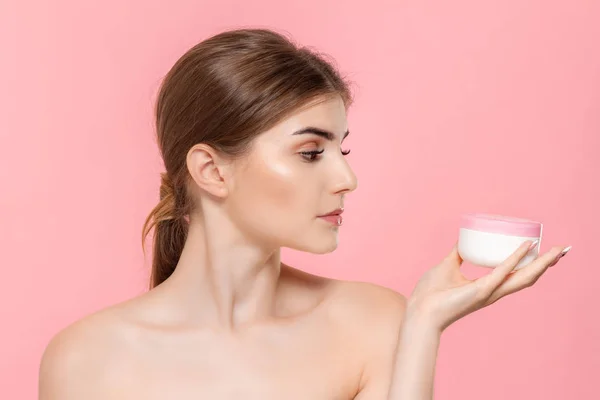 Güzel kız cildi nemlendirmek için krem tüp tutan. Model pembe arka plan üzerinde izole kamera bakar. Güzellik ve sağlık tedavisi kavramı.
