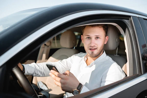 Νέος επιχειρηματίας χαμογελά κοιτάζοντας το κινητό τηλέφωνο κατά την οδήγηση ενός αυτοκινήτου. — Φωτογραφία Αρχείου