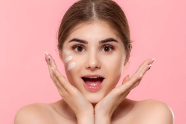 Heyecanlı güzel model pembe arka plan üzerinde izole kamera bakar. Cilde nemlendirici hyaluronik krem uygulanır. Güzellik ve sağlık tedavisi kavramı.