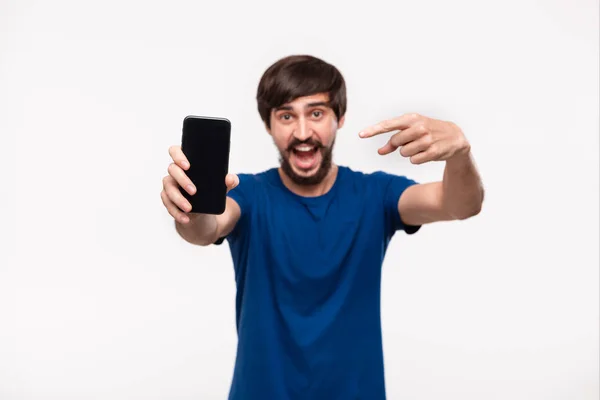 Good looking brunet man in a blue shirt with beard and mostaches shocked looking at the camera pointing at the smartphone standing isolated over white background. Emotion and gesture of surprise. — Stock Photo, Image