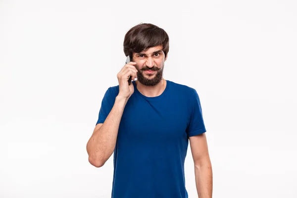 青いシャツにあごひげを生やし、白い背景の上に立ってスマートフォンで話すほとんどの痛みを持つ不愉快なハンサムなブルネットの男。コミュニケーションの概念. — ストック写真
