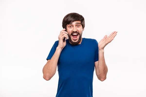 Grazioso uomo bruno in camicia blu con barba e baffi scioccato tenendo smartphone guardando la fotocamera in piedi isolato su sfondo bianco. Emozione di sorpresa. Notizie inaspettate dal telefono . — Foto Stock