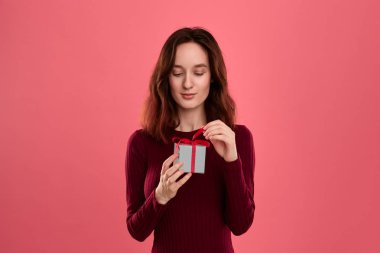 Heyecanlı güzel esmer kız hediye koyu pembe arka plan üzerinde izole ayakta ve kameraya gülümseyerek bir kurdele ile mevcut kutusunda paketlenmiş açılır. Özel etkinliği kutluyoruz.