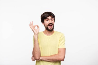 Yakışıklı brunet sakallı adam sarı gömlek bıyıklı ok işareti gösteren beyaz arka plan üzerinde izole ayakta. Tamam hareketi.